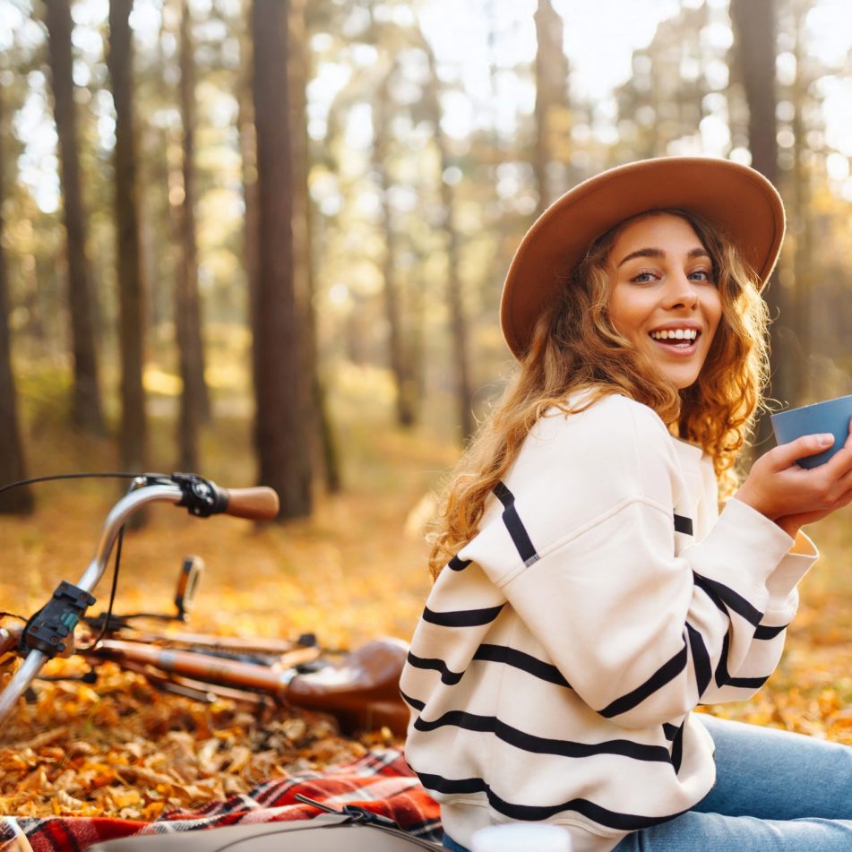 Frau mit Wollpullover und Hut sitzt im Wald auf Laub und hatte eine Tasse in der Hand