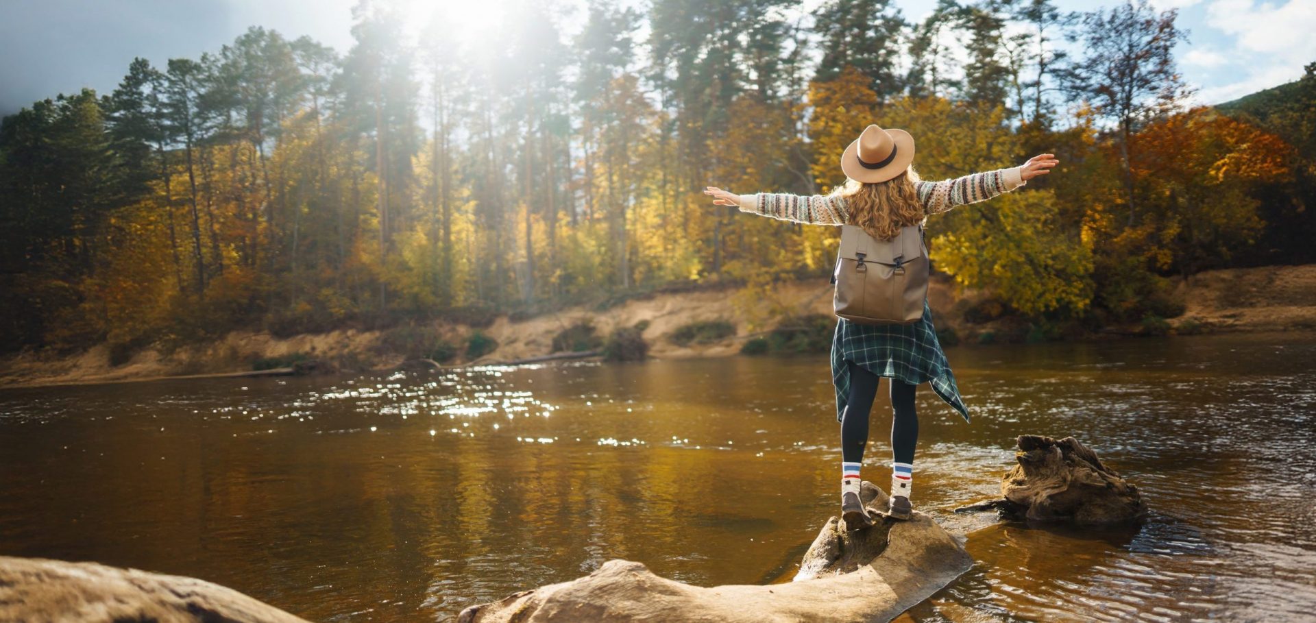 Frau von hinten in Wanderkleidung mit Rucksack und Hut steht auf einem Stein an einem Fluss im Wald und breitet die Arme aus.