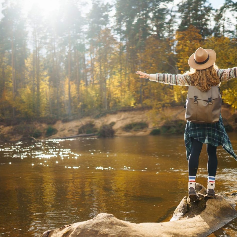 Frau von hinten in Wanderkleidung mit Rucksack und Hut steht auf einem Stein an einem Fluss im Wald und breitet die Arme aus.